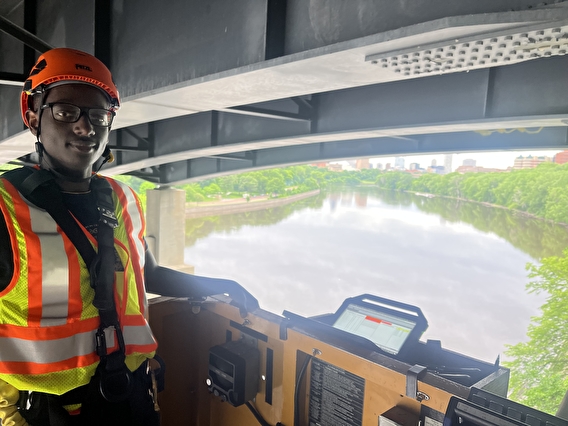 MnDOT intern Ryan Mwangi standing in a snooper beneath a bridge with a river far below.