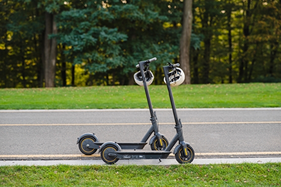 Two e-scooters parked on a sidewalk with a road and trees in the background