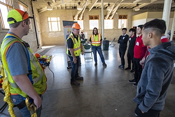 Three workers in yellow safety vests and helmets demostrating protective equipement to a group of high school students