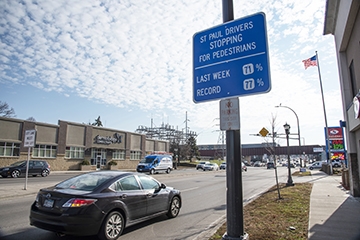 Car next to sign displaying percentage of cars stopping for pedestrians at intersection (77% at time)